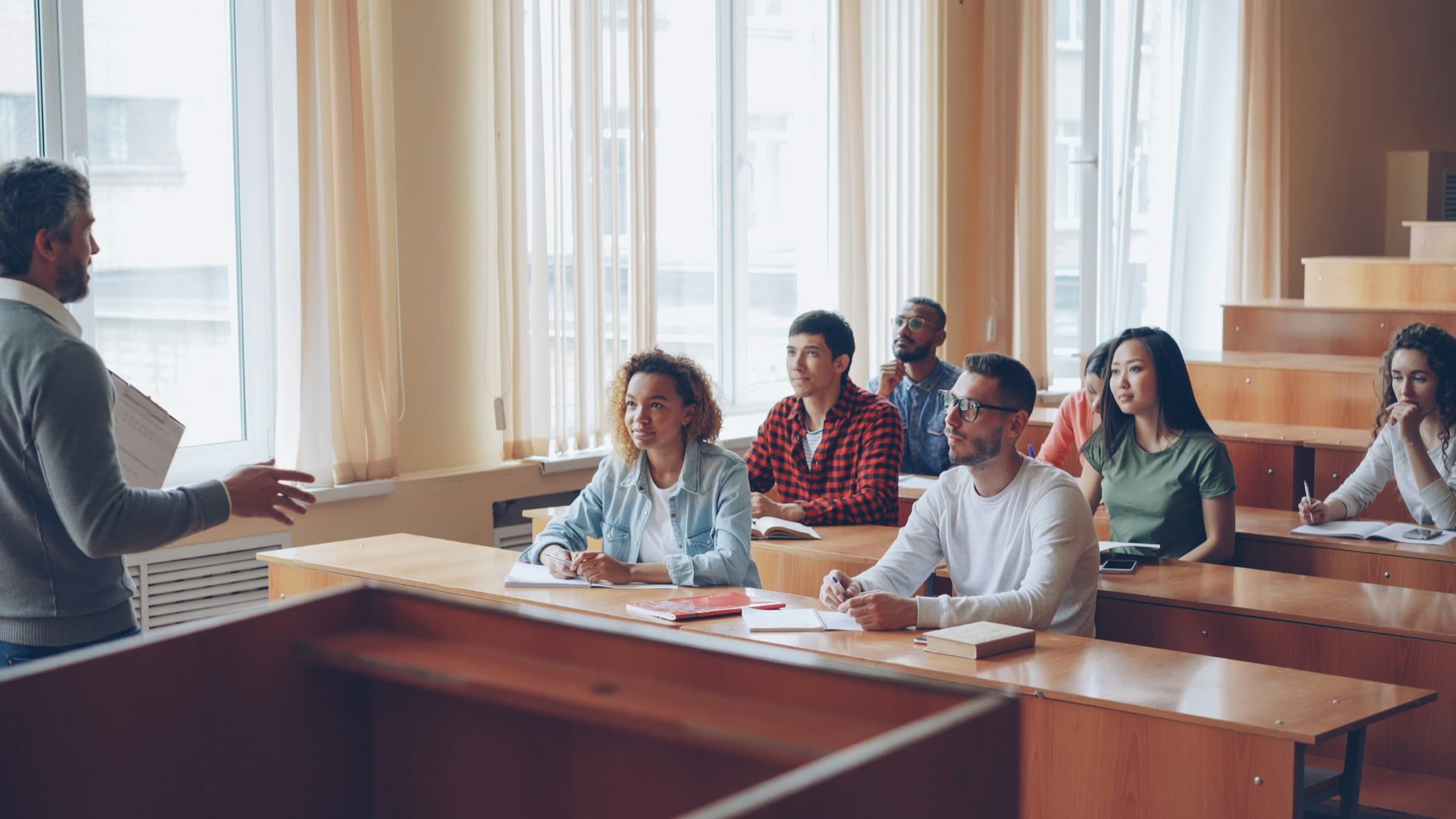 A lecturer gestures to a classroom of attentive, mixed-nationality students seated at wooden desks beside tall sunlit windows
